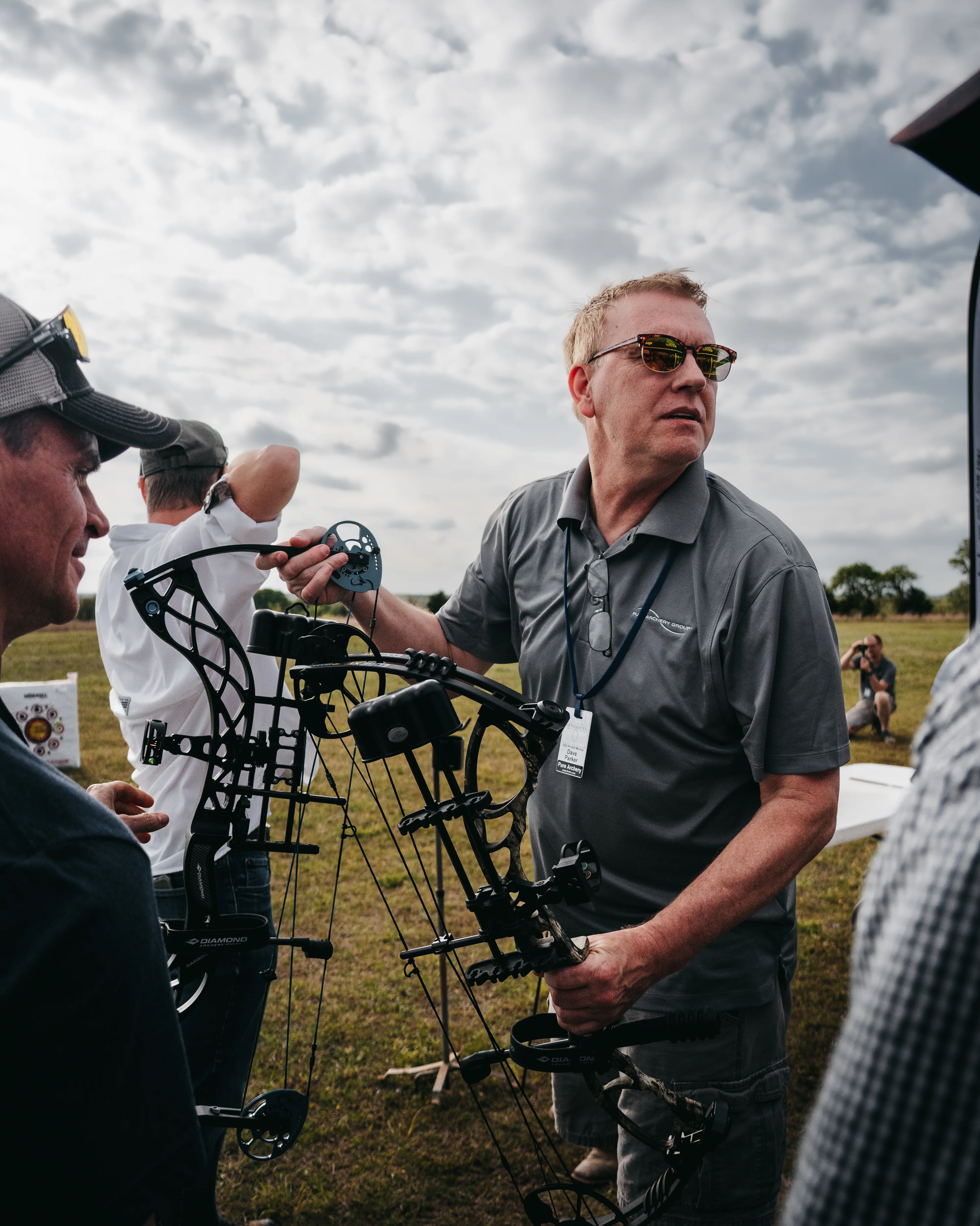 Man holding a compound bow, talking with a group at an outdoor archery event