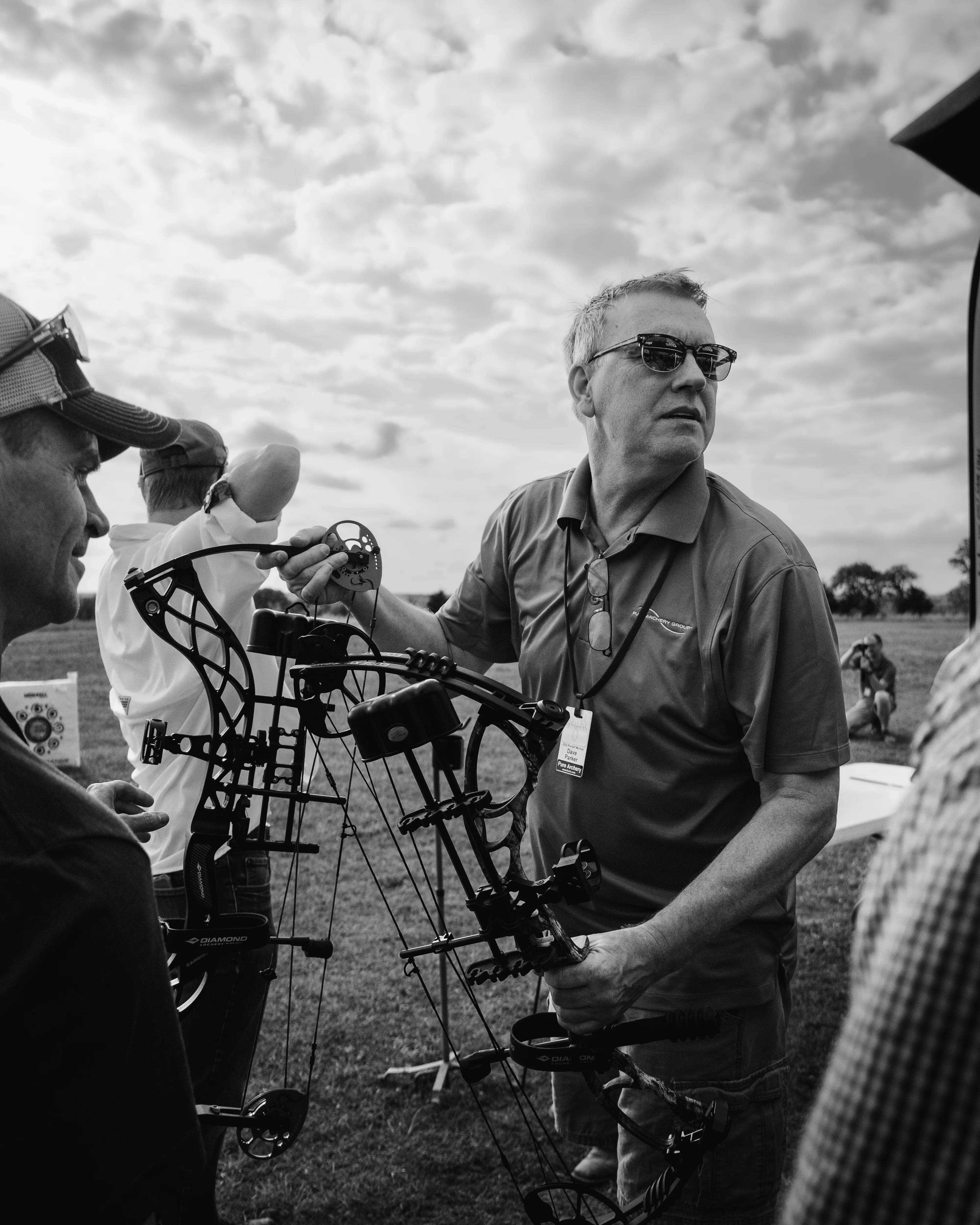 Man holding a compound bow, talking with a group at an outdoor archery event